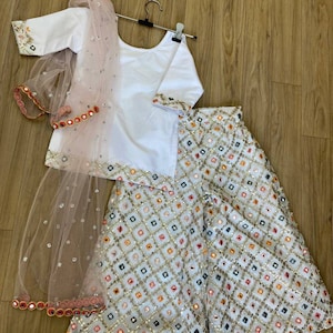 K&ouml;nnte beinhalten: Ein wei&szlig;es und rosafarbenes M&auml;dchen-Outfit mit einem wei&szlig;en Oberteil, einem rosafarbenen Dupatta und einer wei&szlig;en Palazzo-Hose. Die Hose und der Dupatta sind mit Pailletten und Spiegelarbeiten verziert.