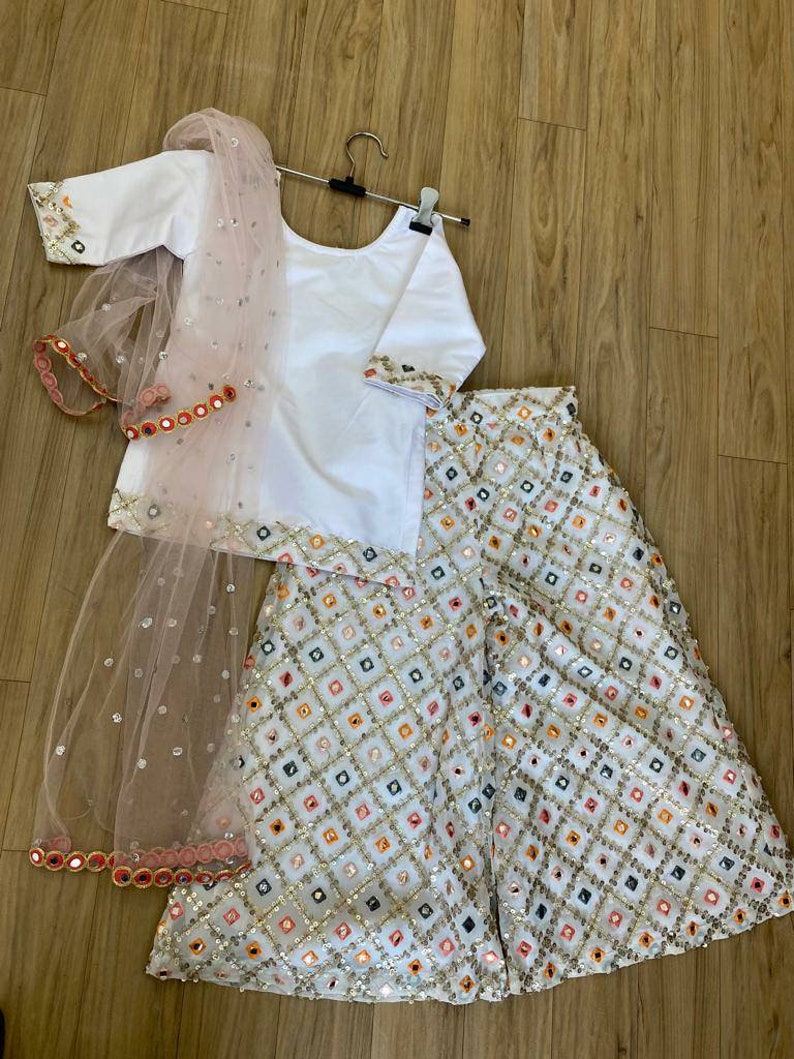 K&ouml;nnte beinhalten: Ein wei&szlig;es und rosafarbenes M&auml;dchen-Outfit mit einem wei&szlig;en Oberteil, einem rosafarbenen Dupatta und einer wei&szlig;en Palazzo-Hose. Die Hose und der Dupatta sind mit Pailletten und Spiegelarbeiten verziert.