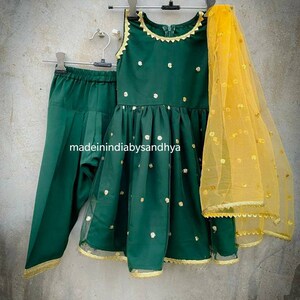 Puede incluir: Un conjunto para ni&ntilde;a en verde y dorado, que incluye un vestido sin mangas, pantalones y una dupatta transparente. El vestido tiene un escote redondo y lunares dorados. Los pantalones son de color verde s&oacute;lido y la dupatta es amarilla con lunares dorados.