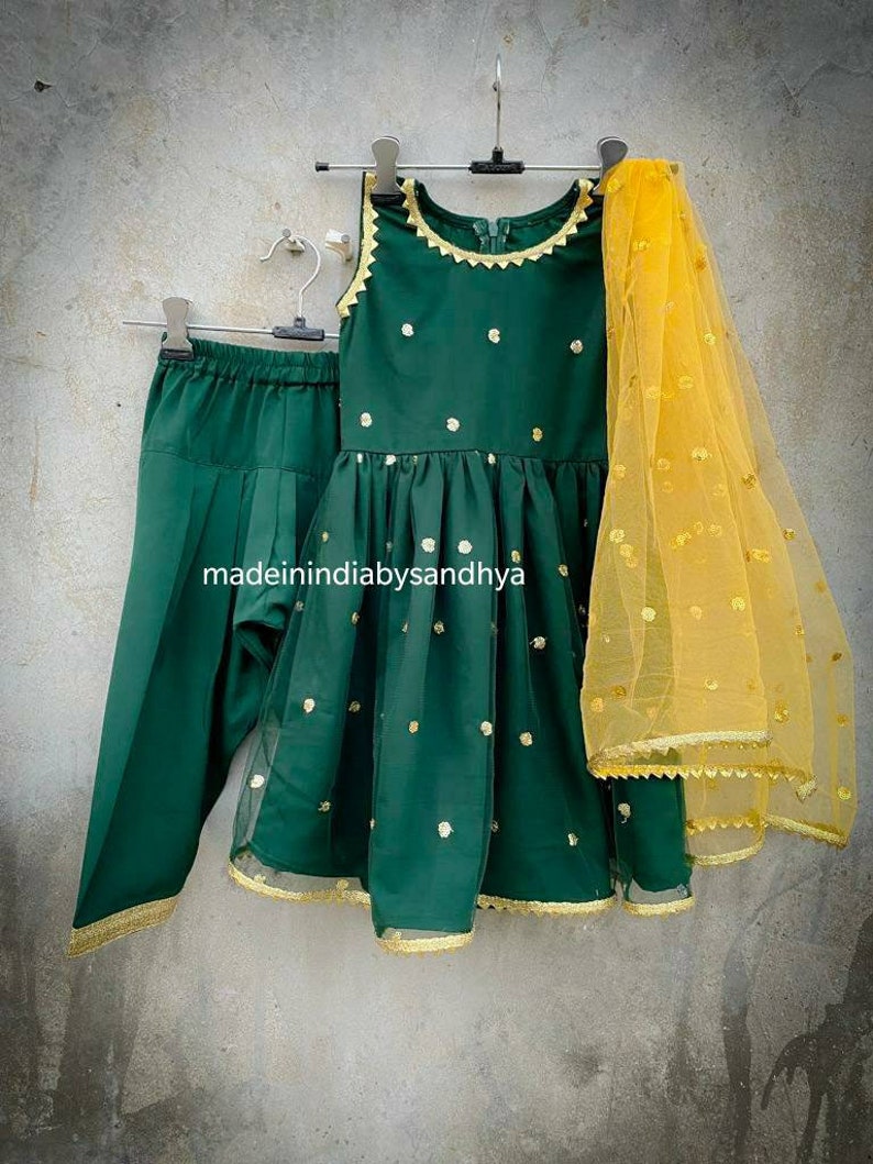 Puede incluir: Un conjunto para ni&ntilde;a en verde y dorado, que incluye un vestido sin mangas, pantalones y una dupatta transparente. El vestido tiene un escote redondo y lunares dorados. Los pantalones son de color verde s&oacute;lido y la dupatta es amarilla con lunares dorados.