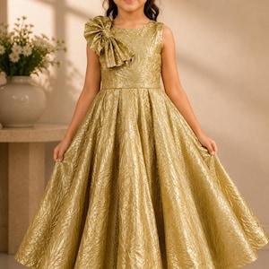 May include: A sleeveless gold dress with a textured, metallic finish and a large, structured bow detail on the right shoulder. The bodice is fitted, transitioning into a full, pleated skirt that flares out, reaching down to the ankles.