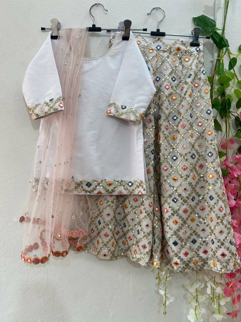 K&ouml;nnte beinhalten: Ein wei&szlig;es und rosafarbenes M&auml;dchen-Outfit mit einem wei&szlig;en Oberteil, einem rosafarbenen Dupatta und einem wei&szlig;en Rock mit bunter Spiegelarbeit.