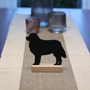 Könnte beinhalten: Schwarze Silhouette eines Hundes, der auf einem Holzsockel steht. Der Hund ist nach rechts gerichtet und hat einen buschigen Schwanz. Der Sockel steht auf einem hellbraunen Tischläufer.