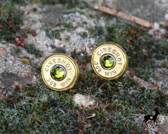 Op de afbeelding: Een paar gouden oorbellen gemaakt van hergebruikte .308 Winchester-kogelhulzen. Elke huls heeft een groene edelsteen in het midden en de woorden "CINE SHOT" en "WIN" gegraveerd rond de rand.