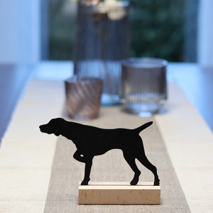 Könnte beinhalten: Schwarze Silhouette eines Hundes, der auf einem Holzsockel steht. Der Hund blickt nach links. Der Sockel steht auf einem hellbraunen Tischläufer.