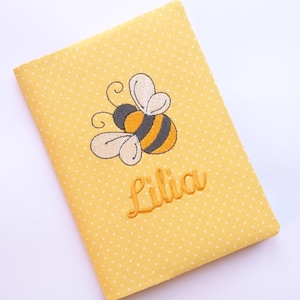 Op de afbeelding: Geel stoffen notitieboekje met witte stippen. Het notitieboekje heeft een geborduurde bij en de naam "Lilia" op de voorkant.