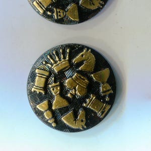 May include: Two round, black buttons with gold chess piece designs. The buttons feature a textured background and various chess pieces, including knights, rooks, and pawns. The buttons are likely for sewing or crafts.