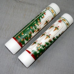 Könnte beinhalten: Zwei weiße Kerzen mit grünen und goldenen Weihnachtsbaum-Designs. Die Kerzen sind mit roten und goldenen Akzenten verziert.