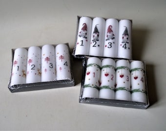 Op de afbeelding: Vier witte kaarsen met verschillende kerstontwerpen. De eerste kaars heeft een rendier en de nummers 1, 2, 3 en 4. De tweede kaars heeft een kerstboom en de nummers 1, 2, 3 en 4. De derde kaars heeft een kerstman en de nummers 1, 2, 3 en 4. De vierde kaars heeft een hart en de nummers 1, 2, 3 en 4.