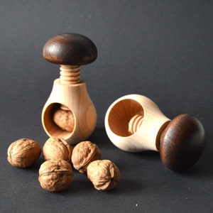 Może przedstawiać: Dwa drewniane łamacze orzechów w kształcie grzybów z ciemnobrązowymi czapkami. Łamacze orzechów znajdują się na czarnym tle z pięcioma orzechami.