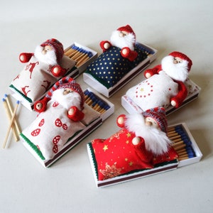Op de afbeelding: Vijf miniatuur kerstman figuren slapen in luciferdoosjes. Elke kerstman draagt een rood-witte muts en een rood-witte deken. De luciferdoosjes zijn versierd met verschillende patronen, waaronder sterren, rendieren en paddenstoelen.