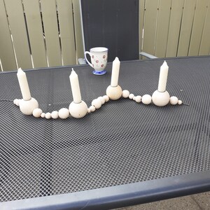 Puede incluir: Un portavelas de madera con una cadena de cuentas de madera. Hay cuatro velas blancas en el portavelas. El portavelas est&aacute; sobre una mesa negra con un patr&oacute;n de rejilla.