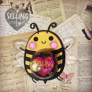 Puede incluir: Una abeja amarilla y negra con una burbuja en forma de corazón que dice "SELLING WITH LOVE" en su interior. La abeja tiene una cara sonriente y mejillas rosadas. La abeja está sobre un fondo de papel viejo con escritura.
