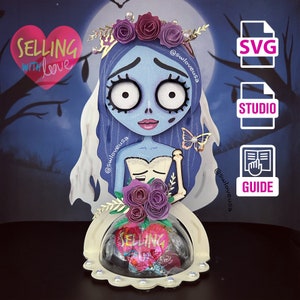 May include: A layered paper art piece featuring a blue-skinned bride with a floral crown and veil. The artwork includes a clear dome with candy and the text "SELLING WITH love" on a heart. Includes SVG, Studio, and Guide icons.