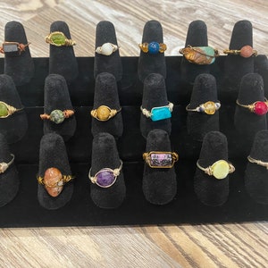 Może przedstawiać: Kolekcja 20 pierścionków owiniętych drutem z różnymi kamieniami szlachetnymi, w tym zielonym, niebieskim, fioletowym, różowym i brązowym. Pierścionki są prezentowane na czarnym aksamitnym stojaku na pierścionki.