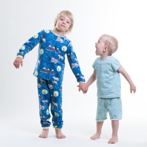 Op de afbeelding: Twee kinderen in pyjama's, een draagt een blauwe pyjama met een berg- en dierenprint, de ander draagt een lichtblauwe pyjama met een witte en lichtblauwe print. Ze houden elkaars hand vast en lachen.