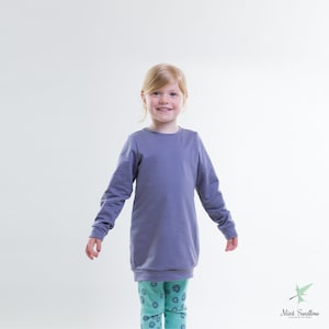 Könnte beinhalten: Ein junges Kind trägt ein langärmliges, lilafarbenes Sweatshirt mit Rundhalsausschnitt. Das Sweatshirt ist im Tunikastil und wird mit gemusterten Leggings getragen.