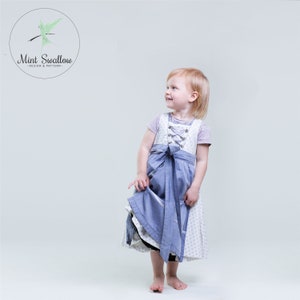 May include: A young girl wearing a white and blue dress with a blue apron. The dress has a white lace trim and a white bow at the waist. The girl is smiling and looking to the side.