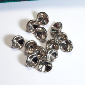 Könnte beinhalten: Eine Ansammlung von elf kleinen, kreisförmigen Metallknöpfen in Silberoptik. Jeder Knopf weist eine glatte, reflektierende, kuppelförmige Oberfläche mit einem erhöhten Rand auf, was für ein poliertes metallisches Aussehen sorgt. Die Knöpfe liegen locker zusammen.