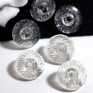 Könnte beinhalten: Sechs durchsichtige Glasknöpfe mit einem erhabenen, strukturierten Design. Die Knöpfe haben eine kleine, durchsichtige Mitte mit einem erhabenen Kreis in der Mitte.