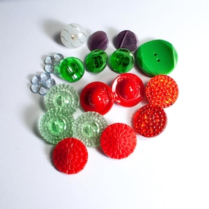Könnte beinhalten: Eine Sammlung bunter Knöpfe in verschiedenen Formen und Größen. Die Knöpfe umfassen transparente, grüne, rote und violette Optionen. Einige Knöpfe haben strukturierte Oberflächen, während andere glatt sind. Einige Knöpfe haben dekorative Elemente.