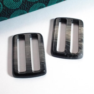 Könnte beinhalten: Zwei schwarze und weiße Kunststoff-Schnallenrutschen. Die Schnallen sind rechteckig mit zwei parallelen Schlitzen in der Mitte.