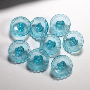 Op de afbeelding: Een verzameling van acht heldere, lichtblauwe glazen knopen. Elke knoop heeft een bloemvormig midden en een decoratieve, geribbelde rand. De ronde knopen hebben een diameter van ongeveer 2,5 cm.