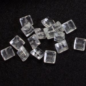 Könnte beinhalten: Eine Ansammlung klarer, quadratischer Perlen mit facettiertem Design. Die Perlen sind auf einer dunklen Oberfläche verstreut, was ihre Transparenz und geometrische Form hervorhebt. Diese Perlen werden wahrscheinlich für die Schmuckherstellung oder das Basteln verwendet.