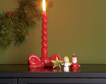 Könnte beinhalten: Ein roter Kerzenhalter aus Keramik in Schleifenform mit einer brennenden, gedrehten roten Stabkerze. Daneben stehen eine kleine goldene Sternfigur, ein Miniatur-Engel mit gelbem Haar und eine winzige Weihnachtsmannfigur mit rotem Anzug.