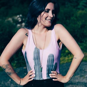May include: A light pink bodysuit with a cactus print. The sleeveless one-piece garment features a scoop neck and is paired with black pants. A gold necklace with a green pendant is worn.