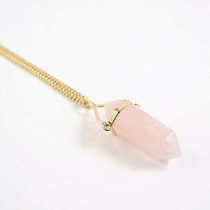 Puede incluir: Un collar de cadena dorada con un colgante de cristal de cuarzo rosa. El cristal tiene forma de punta y est&aacute; sujeto por un alambre dorado.