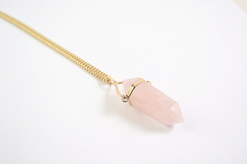 Puede incluir: Un collar de cadena dorada con un colgante de cristal de cuarzo rosa. El cristal tiene forma de punta y est&aacute; sujeto por un alambre dorado.