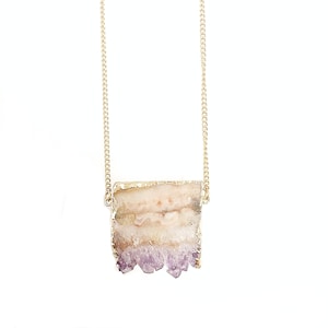 Pode incluir: Colar com corrente dourada e pingente retangular. O pingente é uma pedra natural com borda dourada, exibindo camadas de branco, bege e roxo. O colar é exibido em um fundo branco.