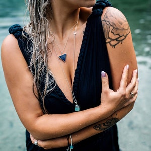 Op de afbeelding: Een vrouw die een zwarte jurk draagt met een diep decolleté en een zilveren driehoekige ketting. Ze heeft een tatoeage op haar schouder van een roos met ranken.