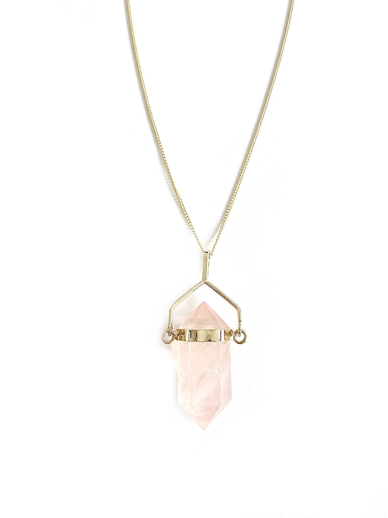 Puede incluir: Un collar de cadena dorada con un colgante de cristal de cuarzo rosa. El cristal es una punta doblemente terminada y est&aacute; suspendido de un marco de alambre dorado.