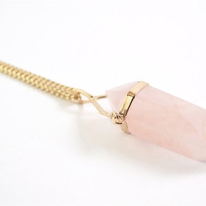 Puede incluir: Un collar de cadena dorada con un colgante de cristal de cuarzo rosa. El cristal tiene forma de punta y est&aacute; engastado en un bisel dorado.