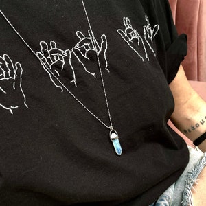 May include: A black t-shirt with white embroidered hand gestures and a silver chain necklace with a blue iridescent crystal pendant.