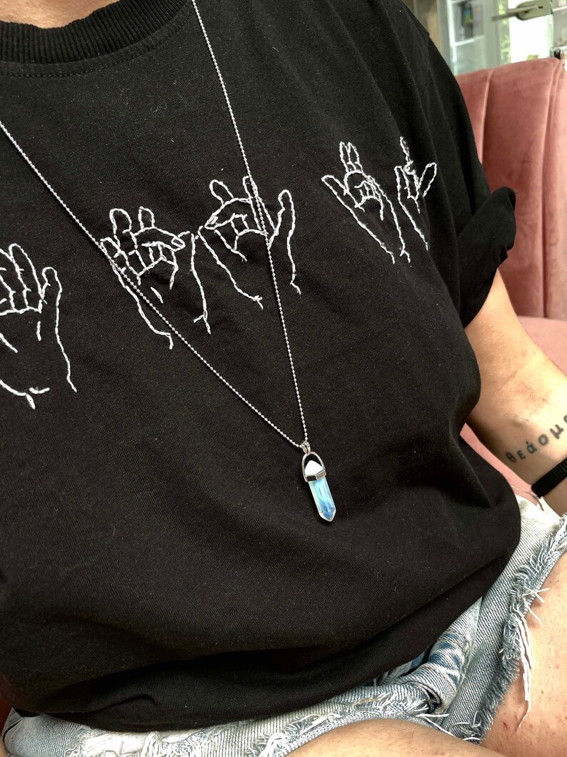 May include: A black t-shirt with white embroidered hand gestures and a silver chain necklace with a blue iridescent crystal pendant.