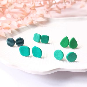 Pode incluir: Uma coleção de brincos de pressão feitos à mão em várias formas e tons de verde e verde-azulado. Os brincos incluem formas redondas, quadradas e em forma de gota, alguns com superfícies texturizadas. Os brincos são exibidos em um prato branco oval.