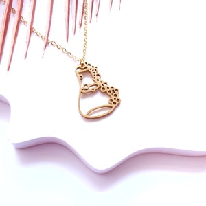 Könnte beinhalten: Eine goldene Halskette mit einem zarten Anhänger, der ein stilisiertes Gesicht mit einem floralen Design zeigt. Der Anhänger ist aus dünnem Metall gefertigt und hat ein ausgeschnittenes Design.