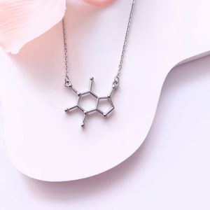 May include: A silver necklace featuring a caffeine molecule pendant. The necklace is displayed on a white surface with a pink flower petal in the background.