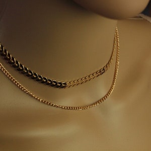 Könnte beinhalten: Zwei goldene Halsketten, die auf einer Schaufensterpuppe übereinander liegen. Die obere Halskette ist eine dicke, klobige Kette mit einem Chevron-Muster. Die untere Halskette ist eine dünnere, zarte Kette.