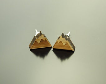 May include: A pair of wooden mountain-shaped earrings with a silver post. The earrings are made of light brown wood with a dark brown mountain design.