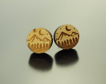 May include: Two wooden stud earrings with a mountain and forest design. Each earring is a small, round disc with a laser-etched landscape featuring mountains, trees, a crescent moon, and stars. The earrings are made of light-colored wood.