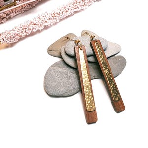 Puede incluir: Pendientes de madera con detalles metálicos dorados. Los pendientes tienen forma rectangular con un panel dorado texturizado en el centro. La madera es de color marrón cálido. Los pendientes se exhiben sobre un lecho de piedras grises.