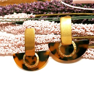 Könnte beinhalten: Goldfarbene Ohrringe mit einem halbkreisförmigen Schildpatt-Design. Die Ohrringe haben einen rechteckigen oberen Teil und einen strukturierten Goldring, der mit der Schildpattbasis verbunden ist. Im Hintergrund sind rosa und lila Blumen zu sehen.