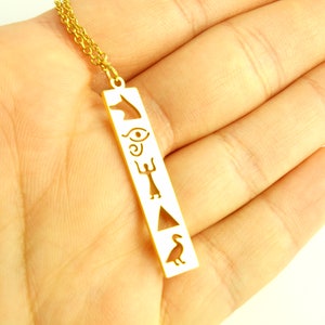 May include: Gold necklace with a pendant featuring an Egyptian-inspired design. The pendant is a rectangular bar with cutouts of an ankh, eye of Horus, and other symbols.