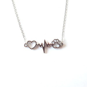 Op de afbeelding: Zilveren ketting met een hart-, hartslag- en pootprintbedel. De ketting ligt op een witte achtergrond.