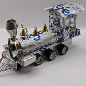 Puede incluir: Un modelo de tren artesanal hecho de latas de aluminio recicladas. El tren presenta una chimenea plateada, campanas doradas y una rejilla frontal detallada. El cuerpo del tren está decorado con gráficos y texto en azul y blanco.