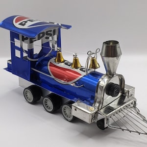May include: A handcrafted train model made from recycled Pepsi cans. The blue and silver train features the Pepsi logo, gold bells, and a silver smokestack. The wheels are black and the train has a metal grill.